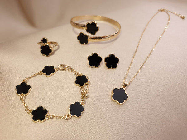 The Set trifoi Negru - auriu 5 piese by Lucky Charm, featuring a gold ring, bracelet, earrings, bangle, and necklace with black clover-shaped charms, is displayed on a soft beige surface.