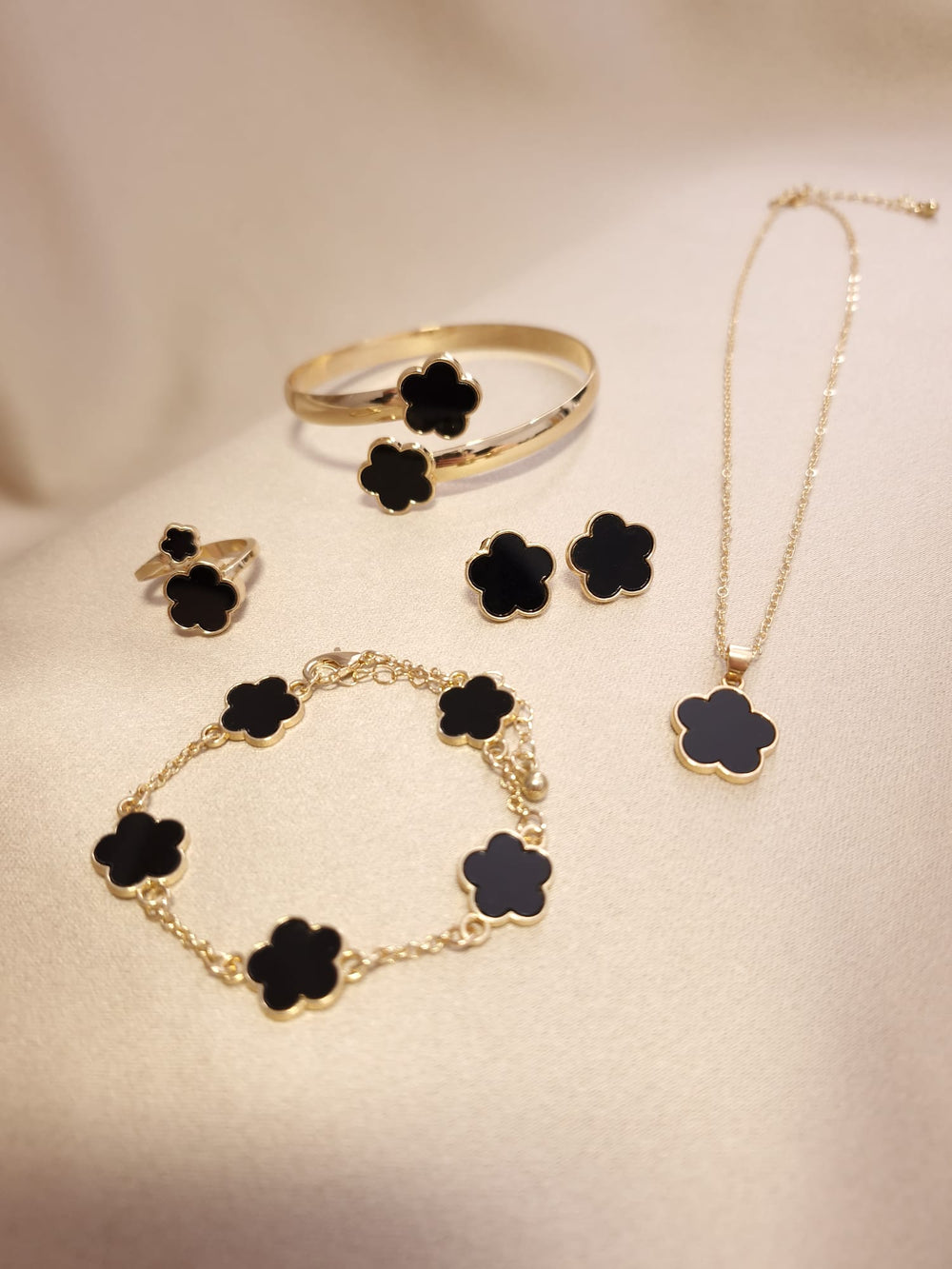 The Lucky Charm Set trifoi Negru - auriu 5 piese features gold jewelry with black clover designs, including a ring, bracelet, bangle, earrings, and necklace displayed on soft beige fabric.
