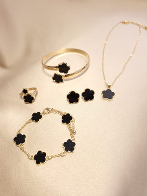 The Lucky Charm Set trifoi Negru - auriu 5 piese features black clover-shaped stones in gold, including a necklace, bracelet, ring, earrings, and bangle, displayed on a light beige surface.
