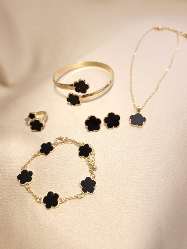 The Lucky Charm Set trifoi Negru - auriu 5 piese features black clover-shaped stones in gold, including a necklace, bracelet, ring, earrings, and bangle, displayed on a light beige surface.
