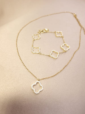 The Lucky Charm Set Trifoi Auriu cu pietre stralucitoare is a gold necklace and bracelet ensemble featuring four-leaf clover-shaped pendants and links, adorned with small clear stones, displayed on a beige surface.