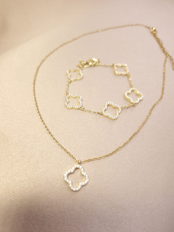 The Lucky Charm Set Trifoi Auriu cu pietre stralucitoare is a gold necklace and bracelet ensemble featuring four-leaf clover-shaped pendants and links, adorned with small clear stones, displayed on a beige surface.