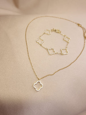 The Set Trifoi Auriu cu pietre stralucitoare by Lucky Charm features a delicate gold necklace and bracelet with diamond-encrusted clover pendants, elegantly displayed on a soft beige fabric background.