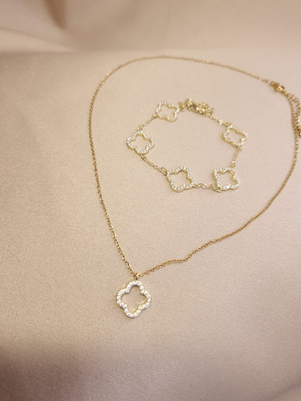 The Set Trifoi Auriu cu pietre stralucitoare by Lucky Charm features a delicate gold necklace and bracelet with diamond-encrusted clover pendants, elegantly displayed on a soft beige fabric background.