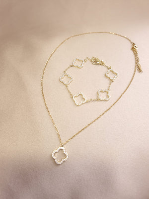 The Set Trifoi Auriu cu pietre stralucitoare by Lucky Charm, consisting of a gold necklace and bracelet with clover-shaped pendants adorned with shiny white stones, is displayed on a light beige fabric background.