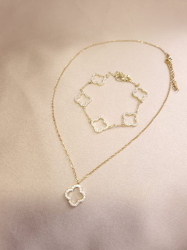 The Set Trifoi Auriu cu pietre stralucitoare by Lucky Charm, consisting of a gold necklace and bracelet with clover-shaped pendants adorned with shiny white stones, is displayed on a light beige fabric background.